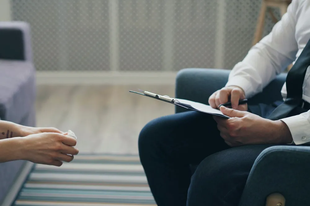 Une séance d’accompagnement vue de côté : les mains d’une personne posées l’une sur l’autre, et celles du praticien qui tient son bloc