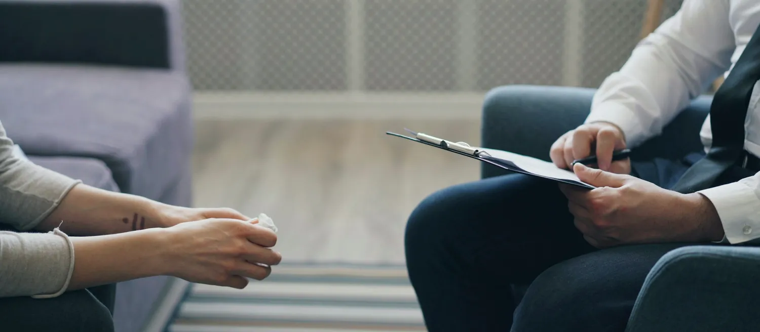 Une séance d’accompagnement vue de côté : les mains d’une personne posées l’une sur l’autre, et celles du praticien qui tient son bloc